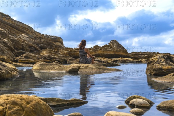 Woman sitting on a rock formation in a coastal landscape, overlooking a tranquil tidal pool, finding a moment of calm and solitude in the natural environment of mount jaizkibel