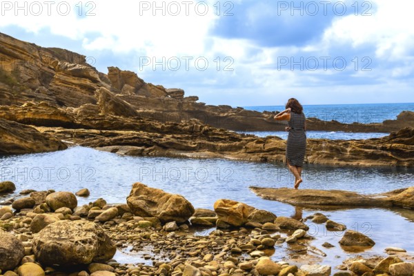 Woman standing barefoot on a rock, observing the peaceful blue sea framed by natural rocky outcrops and geological formations along a serene coastline, finding a moment of solitude