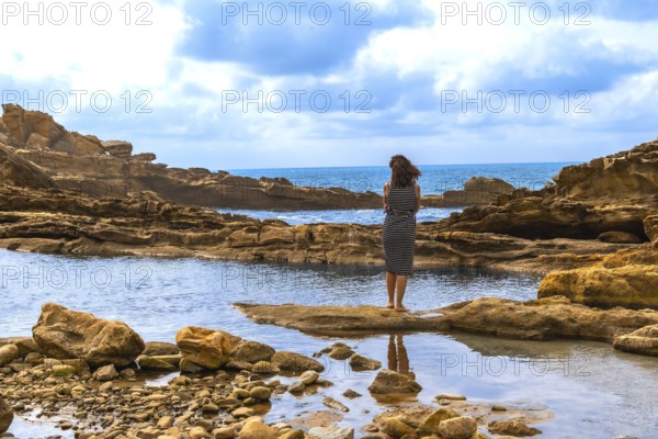 Woman stands barefoot on coastal rock at mount jaizkibel, gazing across the sea while a calm tidal pool reflects sky and shoreline in a serene, contemplative moment
