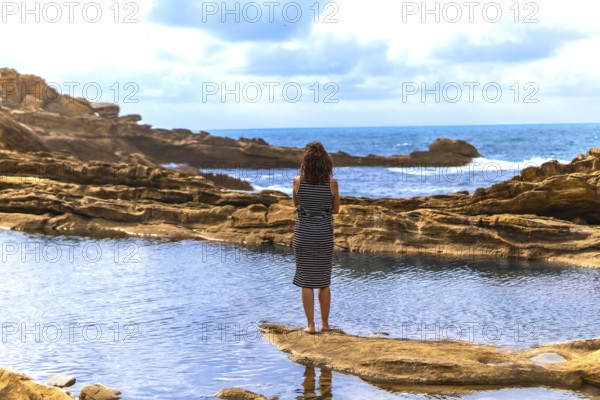 Woman standing barefoot on a rocky outcrop, observing the vast sea and dramatic sky, finding a moment of solitude and contemplation by the natural tide pools