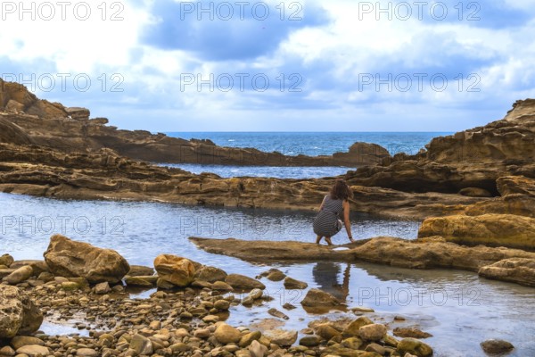 Woman crouching by a rocky tide pool on a rugged coastline, exploring the shallow water with the blue ocean and a cloudy sky in the background, showing nature and adventure