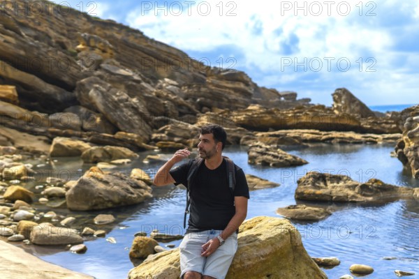 Man with a backpack sitting on a coastal rock, looking to the side and shielding his eyes from the sun, with a rugged rocky beach and a calm water pool stretching behind him under a blue sky