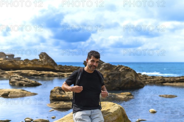 Man with a beard and backpack standing on coastal rocks by the sea, exploring the unique geological formations and vibrant blue sky on mount jaizkibel in basque country, spain
