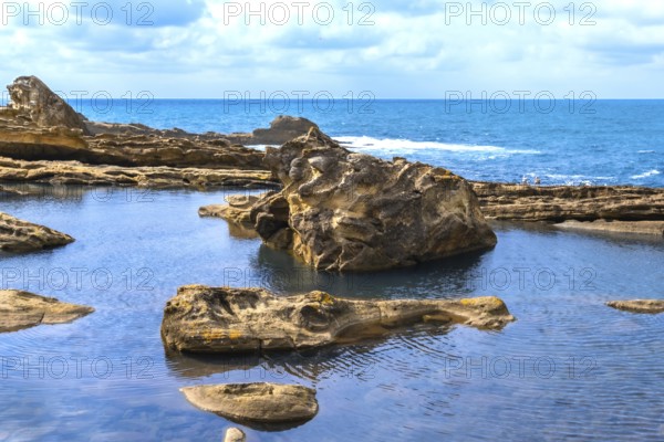 Rocky coastline creating natural tide pools and textured rock formations, showcasing geological erosion with the open ocean and a blue sky in the background