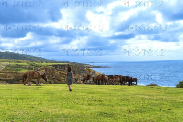 Woman standing on a grassy cliff top observing a herd of wild horses grazing near the edge, with the vast blue ocean and cloudy sky forming a dramatic coastal landscape background