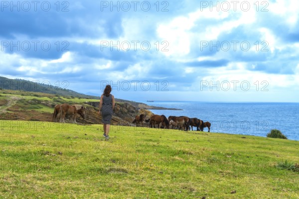 Woman standing on a green cliffside looking at a group of wild horses freely grazing near the blue ocean under a cloudy sky, representing freedom and nature