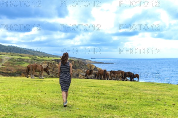 Woman walking along the verdant grass of jaizkibel mountain, observing wild horses grazing freely on a coastal cliff overlooking the vast deep blue atlantic ocean under a cloudy sky