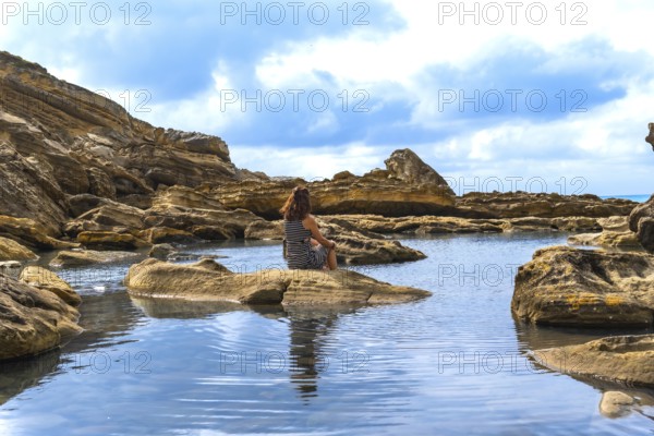 Woman contemplating the serene environment, sitting on a rock by a tranquil ocean rock pool with a calm water surface reflecting the blue sky and textured cliffs