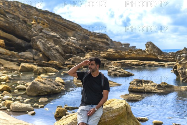 Man in outdoor clothing and backpack exploring a rugged coastal area with geological formations and rock pools, shielding his eyes while looking into the distance on a sunny day