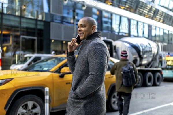 Businessman using a mobile phone on a bustling manhattan street, communicating while cars, including a yellow taxi, pass by high rise buildings, representing urban connectivity and professional life