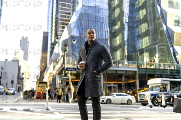 Confident businessman standing on a manhattan street with hand in a modern coat pocket, holding a coffee cup, looking thoughtfully at the urban environment with contemporary buildings