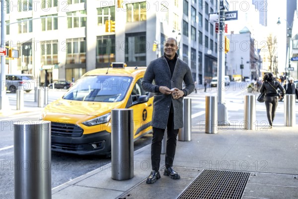 Young businessman stands on a manhattan sidewalk holding a smartphone beside a yellow taxi, smart casual coat signaling confidence during a busy new york commute and urban workday