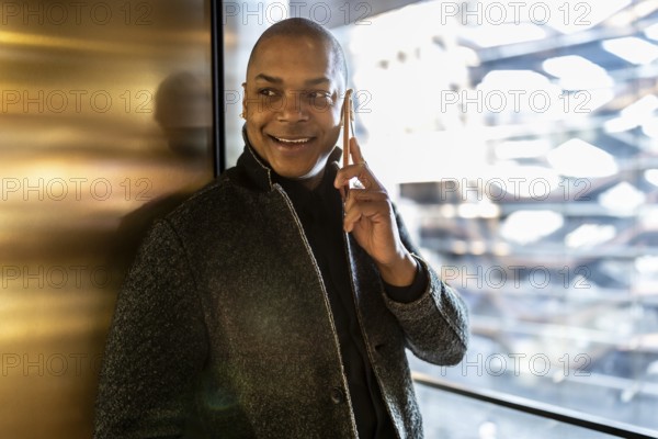 Smiling professional man engaging in a phone conversation, using a smartphone while standing confidently in a modern urban building with bright window light and a reflective wall
