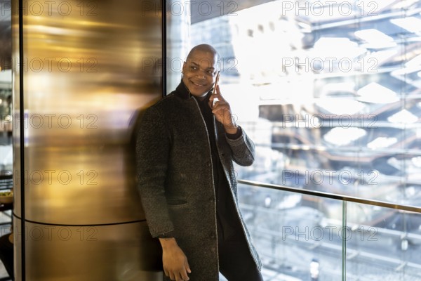 Black businessman standing in a modern building with city views, engaged in a phone call and confidently smiling, representing urban corporate connectivity and success