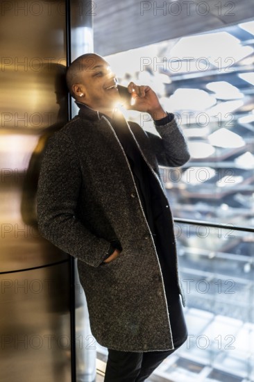 Man in an elegant coat making a phone call, leaning against a metallic wall inside a sunlit elevator with a blurred modern building visible outside, reflecting professionalism and connectivity