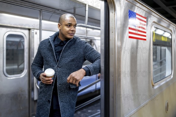 Man standing at a new york subway entrance with a takeaway coffee, looking away while waiting to board urban commuter in professional coat amid bustling transit life