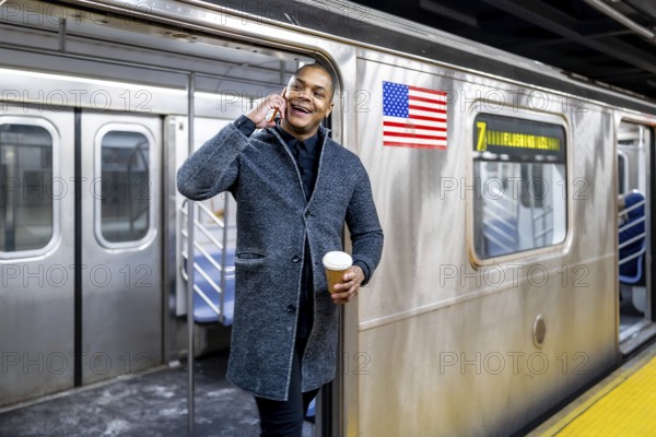 Smiling young professional holding a coffee and talking on his smartphone while stepping off a manhattan subway train, commuting confidently through busy city life