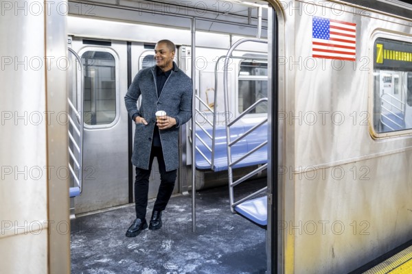 Young businessman standing on a subway car with a coffee cup, looking out through the open door, representing urban commute, travel, and daily city life in new york