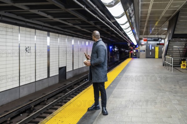 Businessman in a winter coat stands on the yellow safety strip of a subway platform, holding a phone and a coffee cup while waiting for public transport in manhattan, new york city