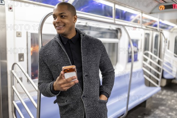 Young african american businessman standing on a subway train in new york city, smiling happily while holding and looking at his mobile phone during an urban commute