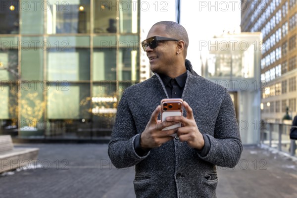 Professional black man smiling in a stylish gray jacket and sunglasses, walking manhattan streets while using a smartphone, confident and connected in an urban business setting