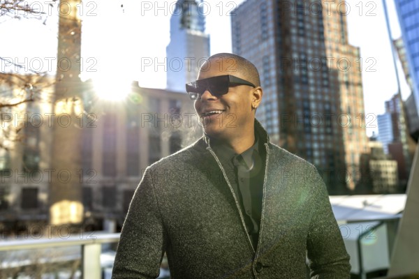 Young man smiling and looking away while wearing dark sunglasses and a gray jacket against a sunny urban backdrop with city buildings, representing a successful lifestyle