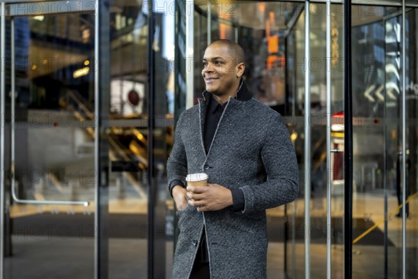 Confident young businessman standing outside a modern office building, holding a disposable coffee cup and looking optimistic, representing urban professional life and success