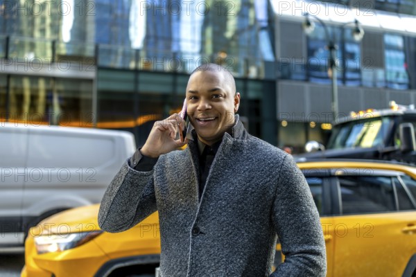 Smiling businessman making a phone call, standing on a busy street in manhattan with cars and modern office buildings in the background, representing communication and urban business
