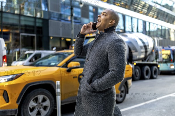 Young professional in a coat making a phone call while walking through a busy manhattan street with yellow taxis and tall buildings in the background, commuting to work