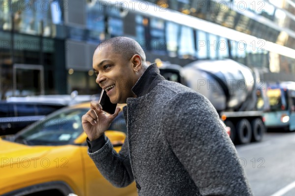 Smiling young executive talks on smartphone while walking a busy manhattan street, surrounded by traffic, yellow taxis and modern office buildings, confident and on the move