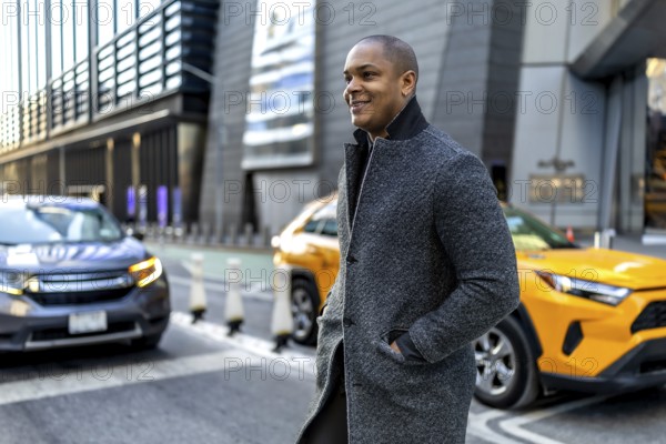 Happy young businessman walking through a busy downtown manhattan street, wearing a stylish gray wool coat, confidently navigating the city scene with a yellow taxi cab passing