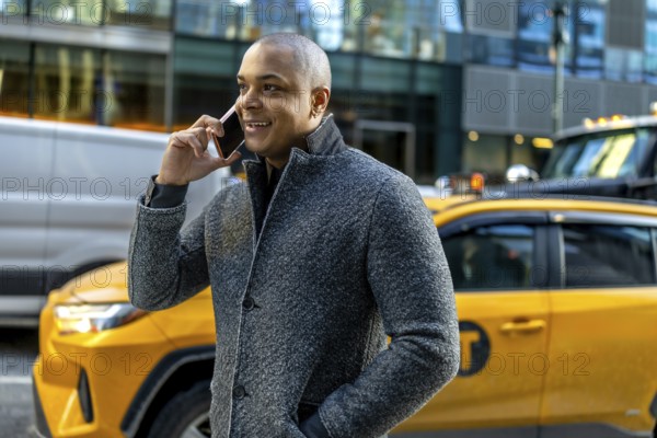 Busy professional man in a winter coat smiling and talking on his smartphone while walking through manhattan with a yellow taxi passing in the busy urban streetscape
