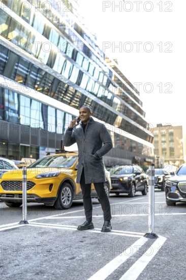 Professional man having a mobile phone conversation on a city street, standing confidently amidst urban traffic with a modern skyscraper in the background