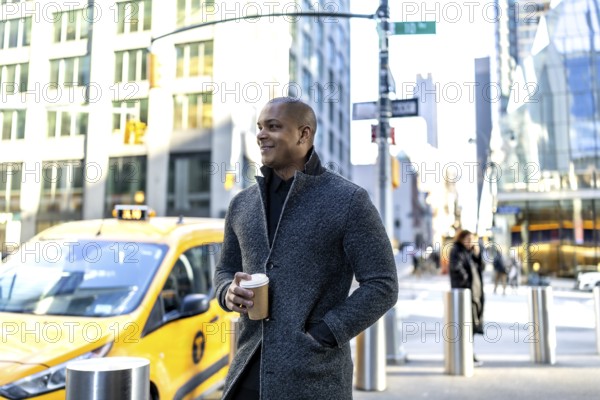 Young man holding a coffee cup, looking thoughtfully while standing on a busy street in manhattan, new york city, with a yellow taxi cab passing by, representing urban lifestyle
