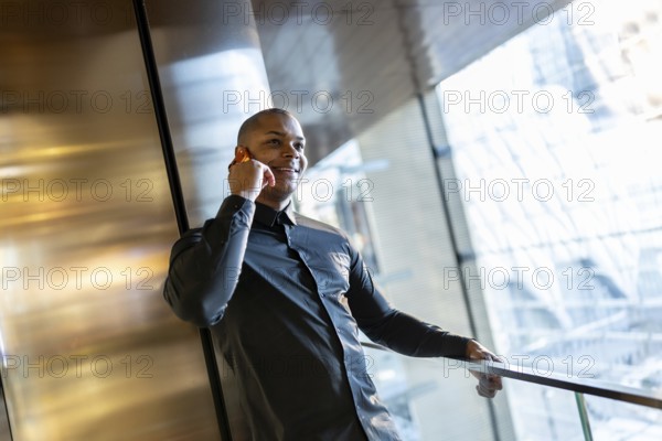 Smiling young professional man is confidently speaking on his cell phone while leaning on a railing inside a sleek urban building, possibly an office or transit hub, conducting business in motion