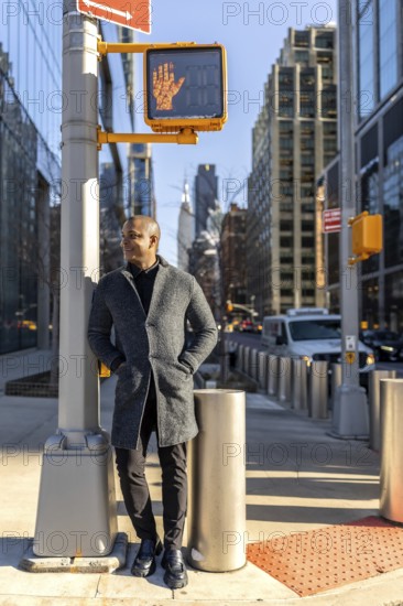 Smiling man standing on a manhattan street at a crosswalk with buildings and traffic in the background, facing a walk signal and representing urban professional life and success