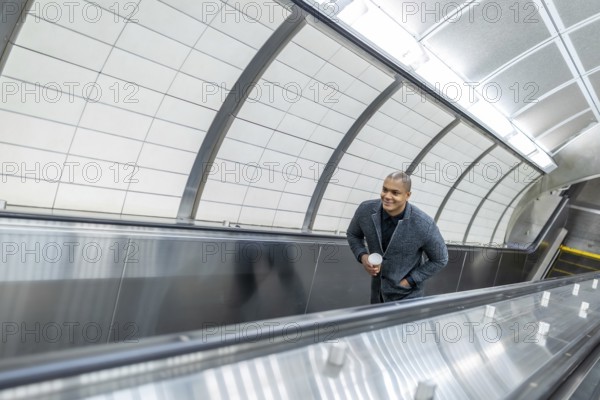 Young businessman ascending a modern escalator in a new york city subway station, holding a coffee cup and looking confidently towards his destination