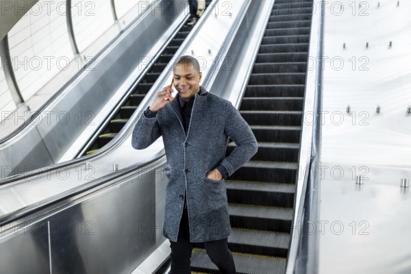 Young man standing on an escalator, holding a cellphone to his ear while smiling, representing urban mobility, communication, and connectivity in a busy metropolitan environment like new york city