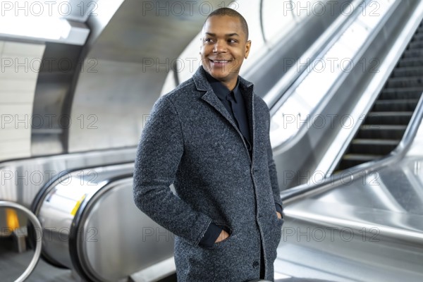 Professional black man smiling, standing confidently on an escalator in a modern subway station, representing urban commute and successful city living