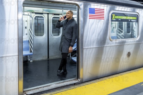 Young man in a stylish coat smiling and talking on his smartphone while standing at the open doors of a subway train on a platform, symbolizing urban connectivity, daily commute, and modern business