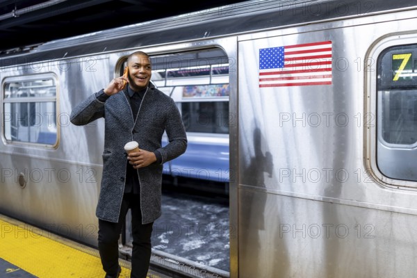 Young businessman commuting in new york city, holding a cup of coffee and having a conversation on his smartphone while walking out of a subway train on the platform