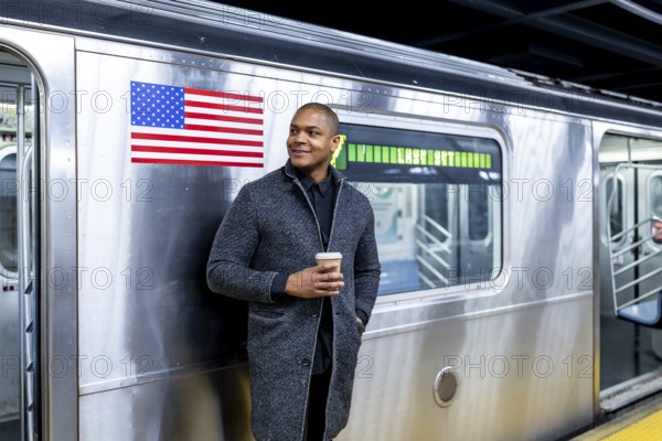 Smiling man standing on a subway platform in manhattan, enjoying a coffee while waiting for public transport, representing urban business travel and daily commute