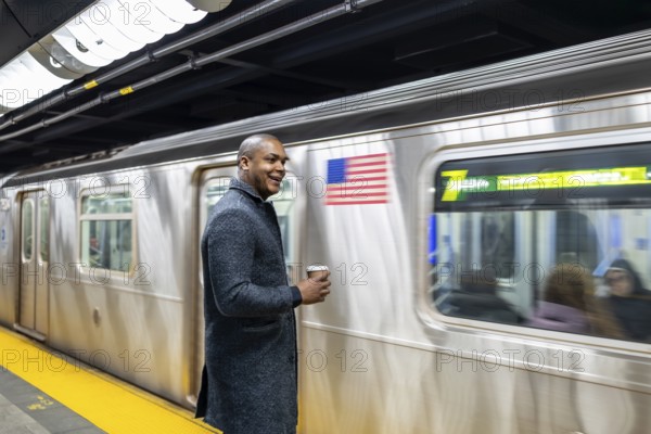 Young businessman standing on a brightly lit subway platform, happily waiting for his train while holding a disposable coffee cup, embracing the urban rush hour