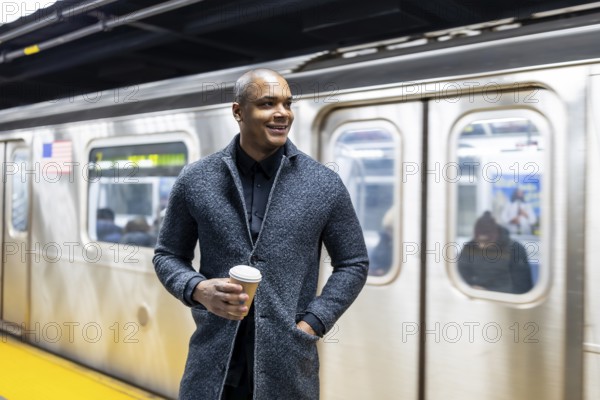 Young businessman standing on a manhattan subway platform with a take out coffee, looking away while waiting for public transport, symbolizing urban commuting and daily city life