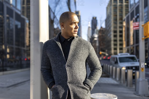 Professional man thinking while standing on a busy new york city street, wearing a stylish gray coat, with modern skyscrapers defining the urban background