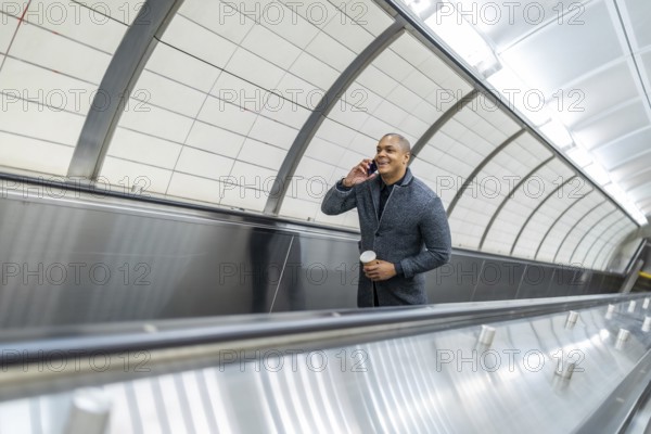 Businessman navigating urban commute on an escalator while engaged in a mobile phone conversation, holding a takeaway coffee, reflecting modern professional life and connectivity in new york city