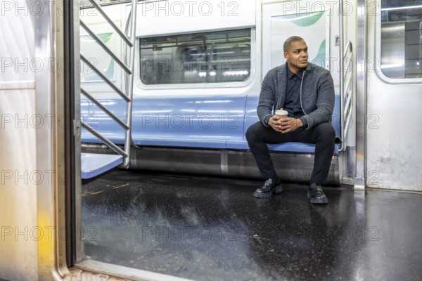 Young man sitting in an empty subway car holding a coffee cup, looking thoughtfully out the window at the urban hustle while traveling to work in manhattan, new york
