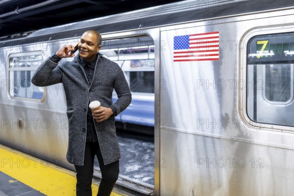 Black businessman standing on a subway platform, talking on a smartphone and holding coffee, commuting in new york city with a train on the 7 line in front of him