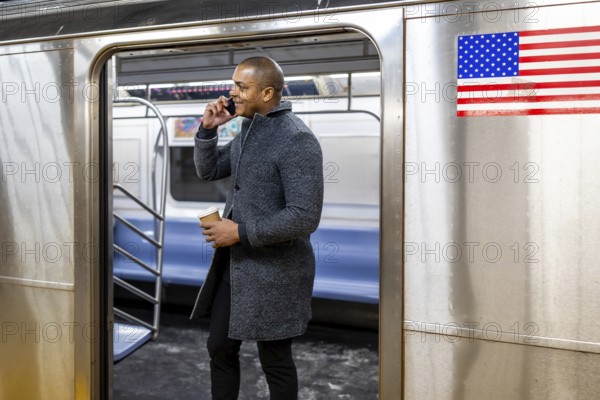 Man holding a coffee cup and having a conversation on his mobile phone, standing inside a new york city subway car with the door open at a platform, representing urban commute and connectivity