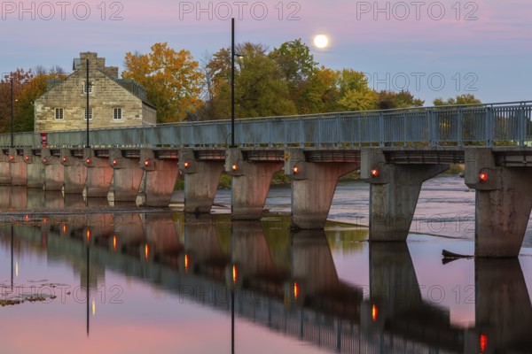 Illuminated Moulin Neuf water flow control dam and walkway over Des Mille-Iles river plus New Mill on Ile des Moulins in autumn at dusk, Old Terrebonne, Quebec, Canada